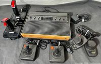 Front view of the Atari 2600 console with six switches and woodgrain front panel, two joystick controllers, two paddle controllers, two additional joystick-style controllers with red buttons, power adapter, and cables arranged around the console.