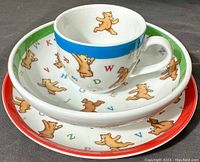Photo of three-piece child's dish set stacked: plate (red rim), bowl (green rim), and cup (blue band), decorated with teddy bears and alphabet letters.