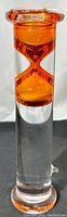 Close-up image of the orange glass hourglass suspended inside a clear glass tube showcasing the clear glass base and rounded top.