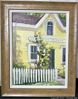 Front view of the framed oil painting showing a yellow bakery building with picket fence and greenery.