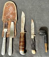 Four vintage cutting tools and briar wood pipe arranged on a gray background: aluminum cutlery set in leather sheath, hunting knife with leather handle, miner's or jack tool, and a textured-handled knife.