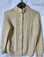 Front view of cream-colored hand knit Aran wool cardigan sweater with spiral buttons, showing knit pattern and button details.