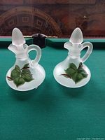 Two frosted glass mini decanters with handles and matching stoppers sitting on a green felt pool table. Both are handpainted with green leaves and subtle detailing.