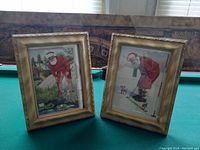 Two framed Santa Claus pictures on a table, one showing Santa golfing and the other skiing, both in gold-colored frames.