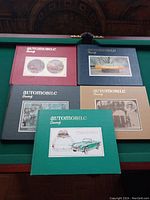 Five automobile-themed hardcover books arranged in two rows on a green surface, each with distinct cover colors and automotive images or text.
