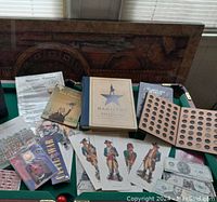 Photo showing Hamilton book, Washington Quarter album, historical postcards, replica US bills, Civil War booklet, and soldier illustrations grouped on a green surface.
