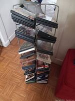 Photo showing a metal vertical CD rack filled with around 37 CDs in jewel cases, positioned near a red couch on a wood floor.