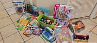 Photo showing assortment of children's toys arranged on floor including puzzles, slime kit, plastic bucket of toys, educational toy cash register, shape sorter, and several small toy figures and books.