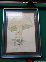 Frontal view of a framed print depicting a child holding a green umbrella standing in a puddle with water ripples around. Blue and gold frame.