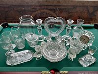 Photo showing a collection of clear glass items including bowls, candle holders, a heart-shaped vase, a glass clock, small dishes, and a decorative tray arranged on a green felt surface.