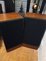 Pair of Realistic floor speakers with wooden trim on the top and bottom with black fabric speaker grills on the front, positioned on wooden floor.