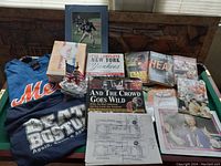 A table with sports memorabilia including New York Mets and 'Beat Boston' T-shirts, various sports books, framed football photo, and printed papers.
