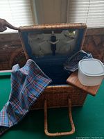 Wicker picnic basket open showing interior fabric lining, two glass bottles, two glasses, plaid tablecloth, small wooden cutting board, and white plastic container with lid.