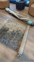 Photo showing two rugs, one small flat rug with intricate traditional medallion and floral pattern, beige tones, fringed edges, and a larger rolled rug with geometric design in neutral tones.