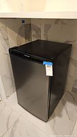 Full view of the black Frigidaire bar fridge placed in corner with tile wall and floor.
