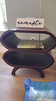 Front view of dark brown rattan side table with oval openings showing a book inside and a "namaste" sign on top, both items not included in lot.