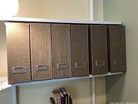 Six brown/neutral fabric magazine boxes lined up on a white shelf, showing their spines with metal label holders.