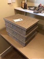 Three stacked rectangular fabric-covered storage boxes with lids in beige woven pattern and metal label holders on front.