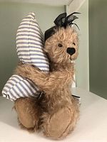 Side profile of light brown mohair bear embracing blue and white striped pillow, showing overall shape and fabric textures.