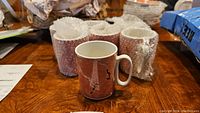 Six Villeroy & Boch Sienna coffee mugs wrapped in bubble wrap on a wooden table, one mug unwrapped and standing upright showing the reddish marble pattern with black design accents.