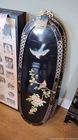 Photo of an oval black lacquer wood wall panel with a blue and orange bird flying and pink blossom branch design, brass mounting hardware visible on top.