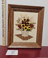 Front view of rectangular framed crewel flower embroidery art with basket of flowers, ruler for scale, and auction lot number.
