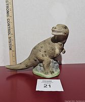 Front side view of the ceramic dinosaur coin bank showing detailed texture and base with rocks and grass.