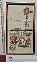 Full front view of framed crewel needlework featuring farm scene with windmill, fence, flowers, and fields. Includes a measuring ruler showing size.