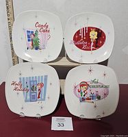 Four white square plates with retro festive Christmas illustrations and text, displayed on stands with visible ruler for scale