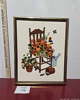 Front view of vintage crewel textile embroidery depicting floral arrangement on a wooden chair with surrounding elements including a watering can and butterfly. Frame in place.