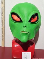 Front view of green rubber alien mask with detailed facial features and measuring scale showing approximate height.