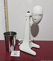 White Hamilton Beach Drink Master mixer stand with stainless steel cup next to a ruler for scale, showing the unit's height and design.