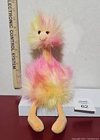 Front view of Jellycat ostrich plush showing its colorful yellow, pink, and peach fur with a measuring ruler alongside indicating size approximately 10 inches tall. The plush has soft legs and black eyes.