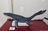 Full body view of large Mosasaurus action figure showing body, tail, and flippers against a neutral background with ruler in the back for size reference.