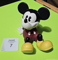 Front view of ceramic Mickey Mouse coin bank showing sitting Mickey with large ears, smiling face, red shorts, white gloves, and yellow shoes.