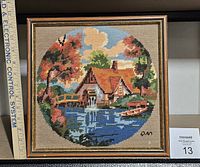 Front view of the framed needlepoint showing the cottage and lake scene with a wooden frame.