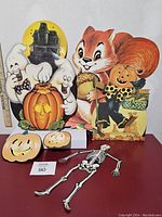 Photo showing a haunted house with two ghosts around a pumpkin, a squirrel holding harvest, scarecrow, two jack-o-lantern faces, and a silver skeleton decoration lying flat.