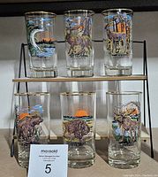 Set of six vintage Canadian wildlife-themed clear glass tumblers with gold rims, arranged on a dual shelf stand, showing all six animal designs including pike, timberwolf, white-tailed deer, grizzly bear, black bear, and moose.