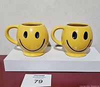 Front view of two yellow ceramic mugs with black smiley faces and handles on the right, displayed side by side.