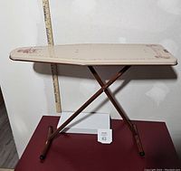 Front and side view of the vintage child's tin litho ironing board set against a wall with a measuring stick for scale, showing the top surface and legs.
