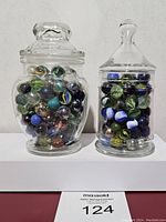 Photo showing two clear glass jars filled with a variety of colorful vintage marbles on a white surface against a plain background.