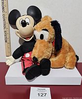 Front view showing the plush Pluto dog sitting next to rubber head Mickey Mouse figure with a ruler behind for size reference.