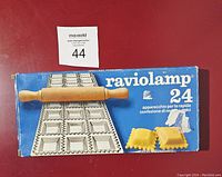 Photo of vintage Raviolamp ravioli maker with original blue box showing product image and rolling pin on top of ravioli mold.