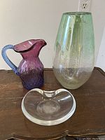 Set of three art glass pieces on wooden surface: purple pitcher with blue handle, clear bowl, greenish vase with bubble texture