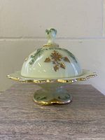 Side front view of the covered butter dish showing the floral hand-painted gold and green design on the lid and scalloped dish edges