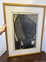 Photo showing the framed abstract etching with geometric shapes in black ink on paper, held at an angle to display the frame and etching details.