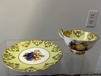 Set of porcelain teacup and saucer with orchard fruit design and gold gilt detail, shown on clear stand.