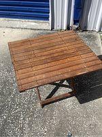 Front angled view of the teak folding table showing the square slatted tabletop and leg base on asphalt surface.