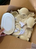 Top view inside cardboard box showing cream-colored china pieces including teapot, creamer or sugar bowl, and serving dish with lids and smaller parts wrapped in cloth.