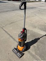 Full view of upright Dyson Ball vacuum cleaner on pavement, showing orange ball base, grey body, and handle.